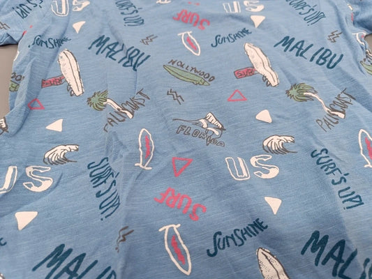 Detailaufnahme des bunten Surfprints mit Malibu-, Sunshine-, Palmen- und Wellenmotiven auf dem hellblauen Jungen-T-Shirt in Größe 98