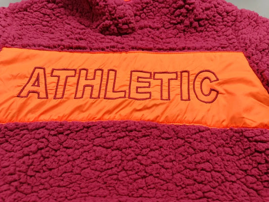 Detailaufnahme der orangefarbenen ATHLETIC-Applikation auf der Fronttasche des pinkfarbenen Teddy-Fleece-Hoodies in Größe 146