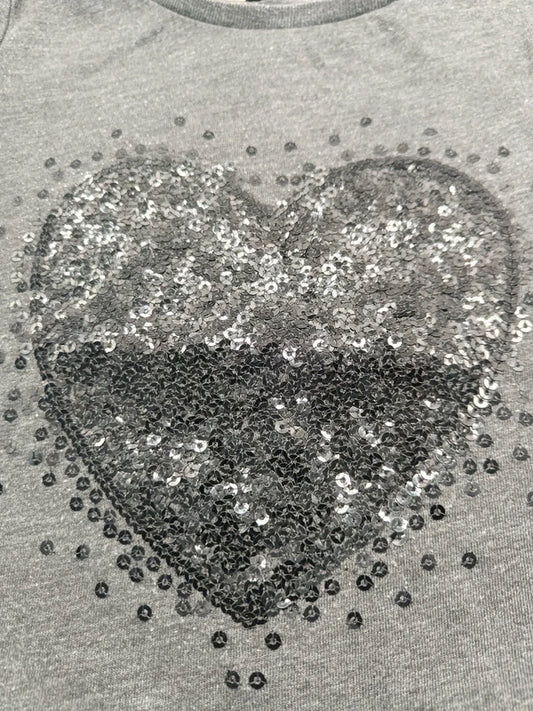 Detailaufnahme des funkelnden silber-schwarzen Paillettenherzes auf dem grauen Mädchen-T-Shirt in Größe 98
